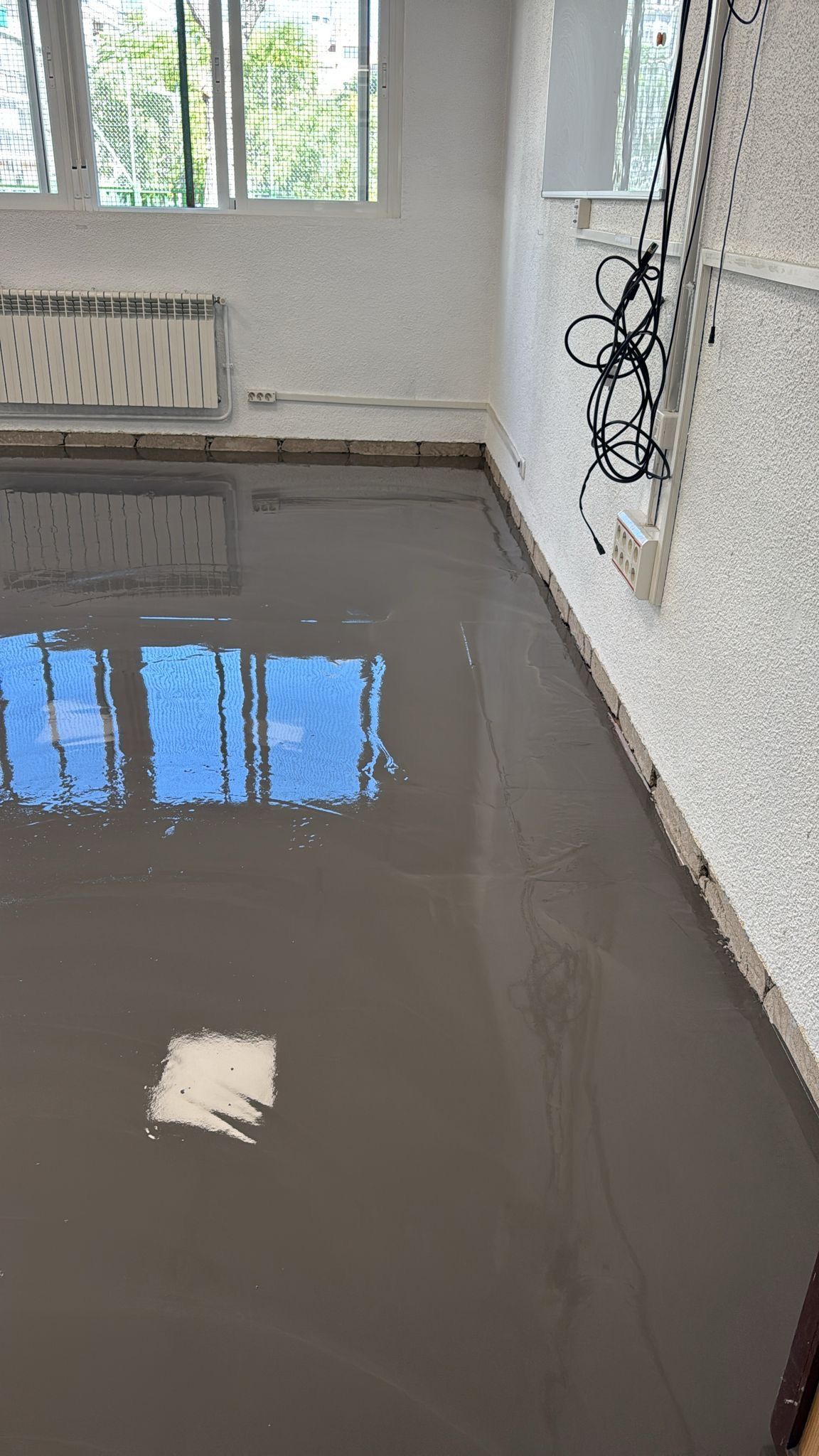 Habitación con suelo gris recién vertido. Las ventanas reflejan el cielo azul. Paredes blancas, radiador y cableado.