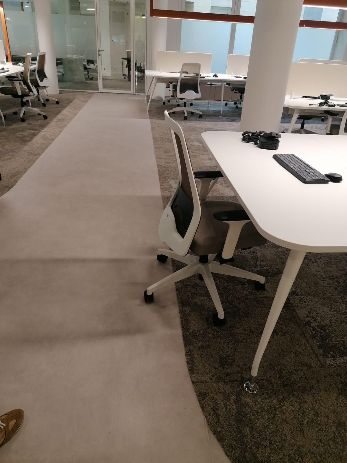 Oficina con escritorios, sillas y alfombra clara. Una silla está junto a un escritorio con teclado.