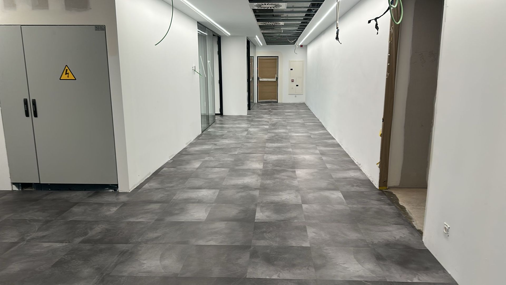 Pasillo largo y vacío con suelo de baldosas grises, paredes blancas y una puerta gris cerrada.