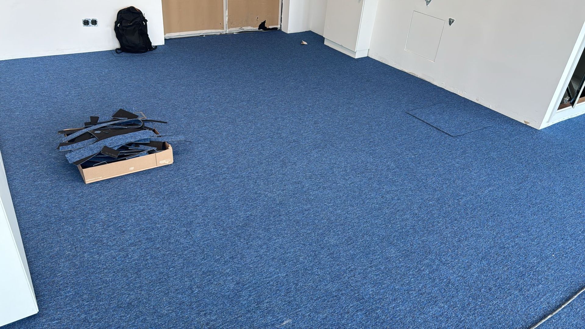 Habitación alfombrada de color azul con una mochila negra y una caja de cartón que contiene artículos negros.