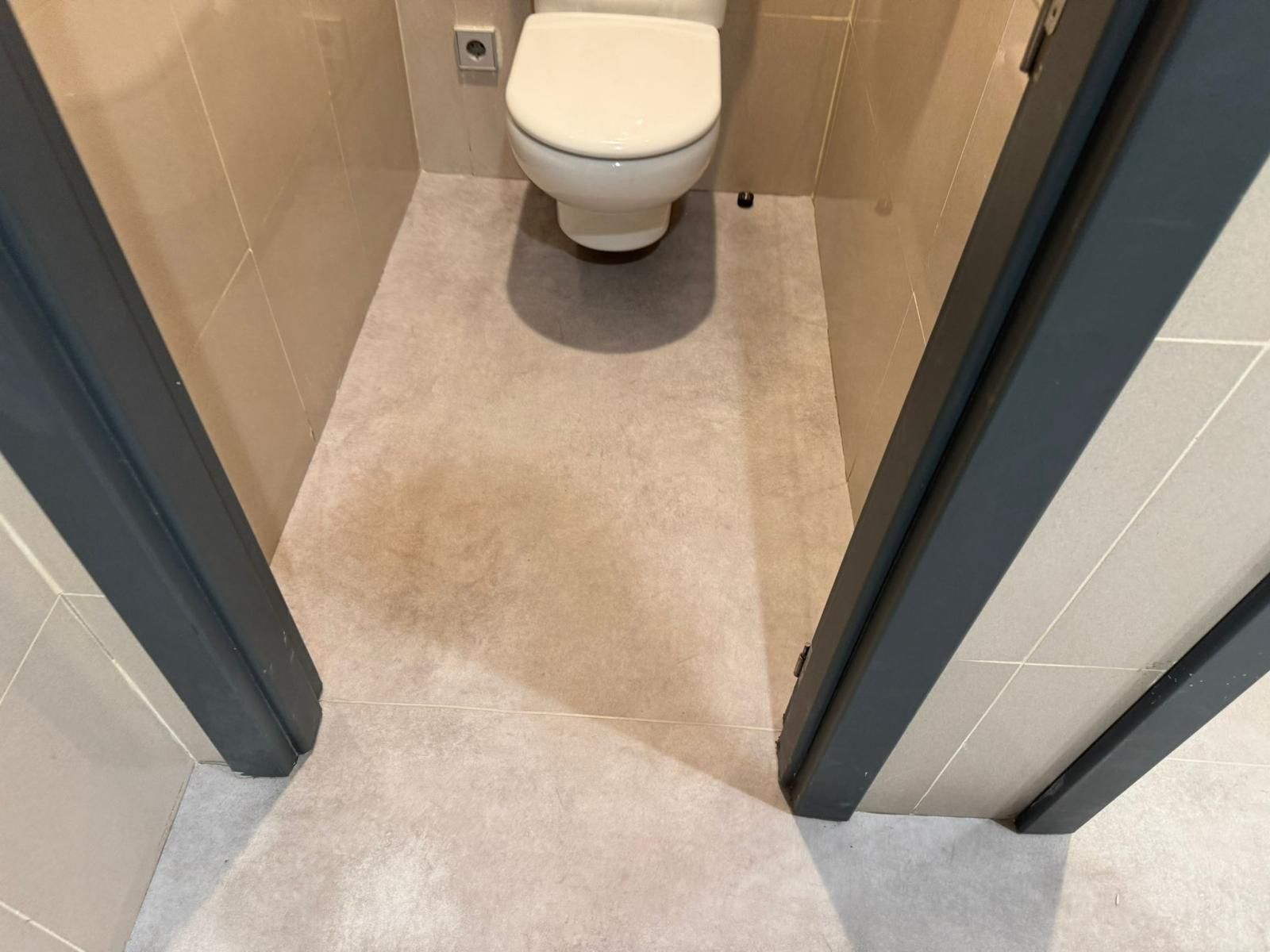 Un baño pequeño con inodoro y suelo gris claro, visto a través de un marco de puerta gris oscuro.