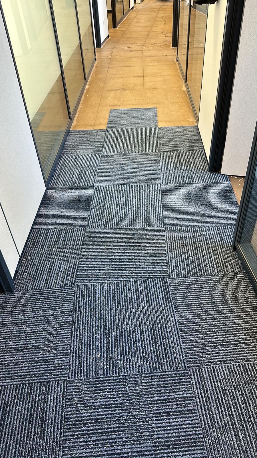 Pasillo de oficina con alfombra estampada y habitaciones con paredes de cristal a ambos lados. Suelo de madera a lo lejos.