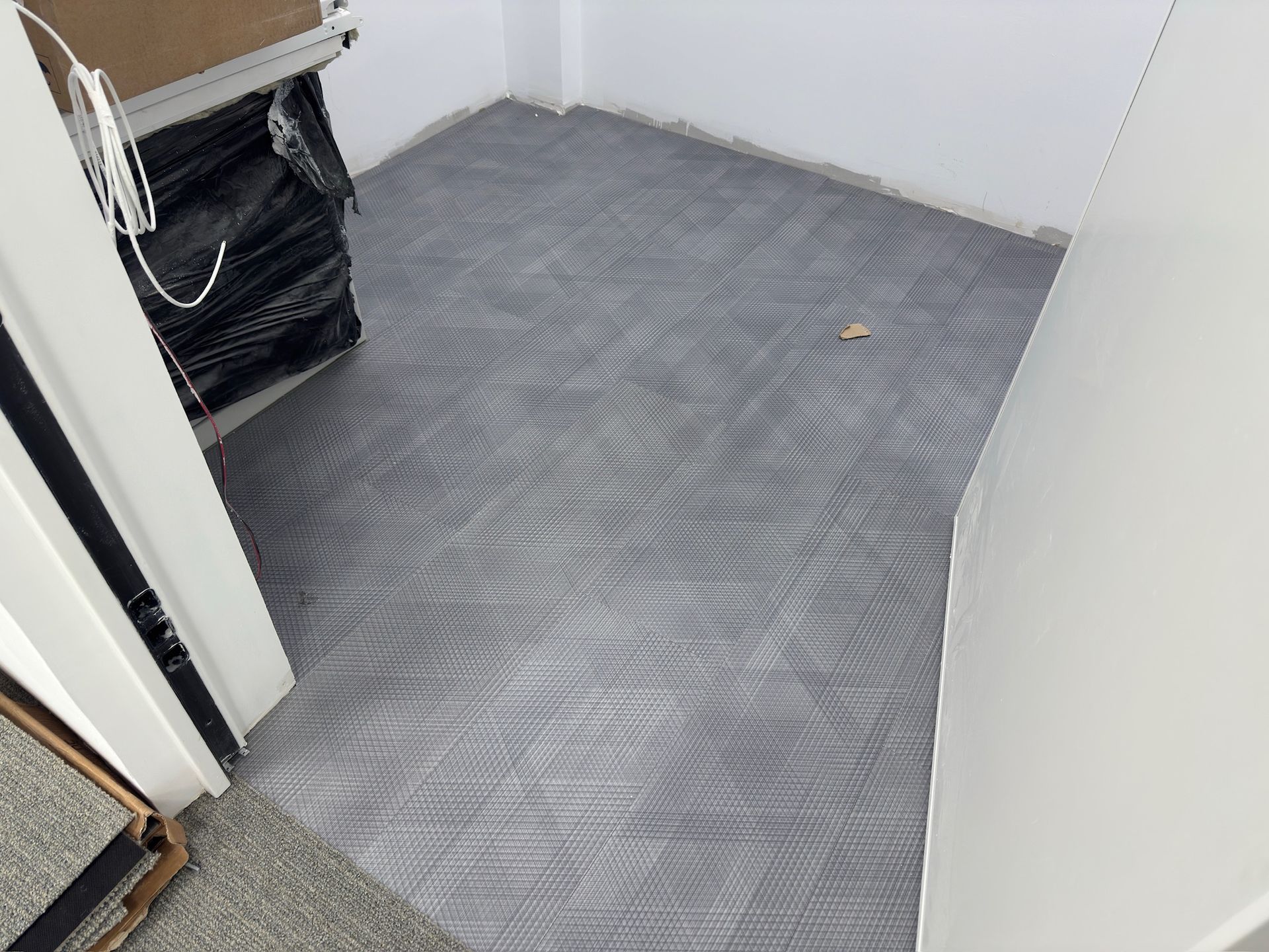 Habitación con baldosas de alfombra gris instaladas; puerta y materiales de piso apilados visibles.