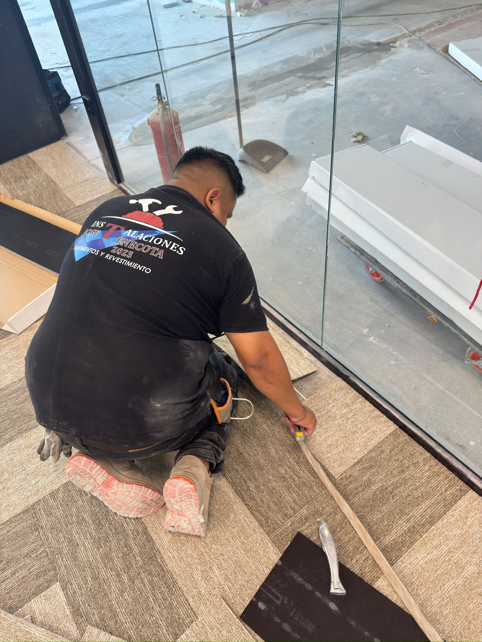 Hombre arrodillado sobre una alfombra estampada, cortando material cerca de una pared de vidrio, con herramientas a su lado.