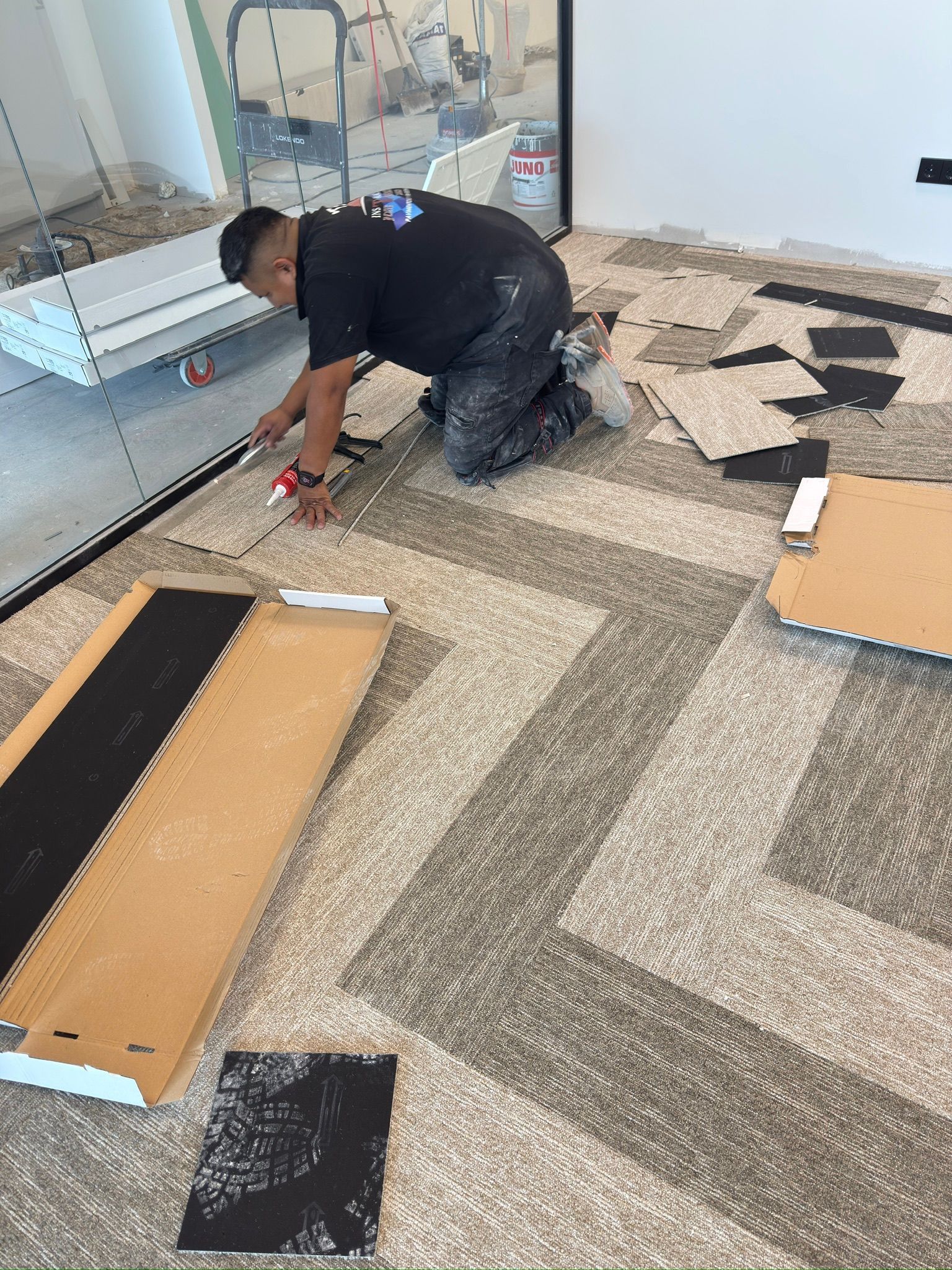 Persona instalando losetas de alfombra usando una herramienta. Las losetas son grises y negras, en una habitación.