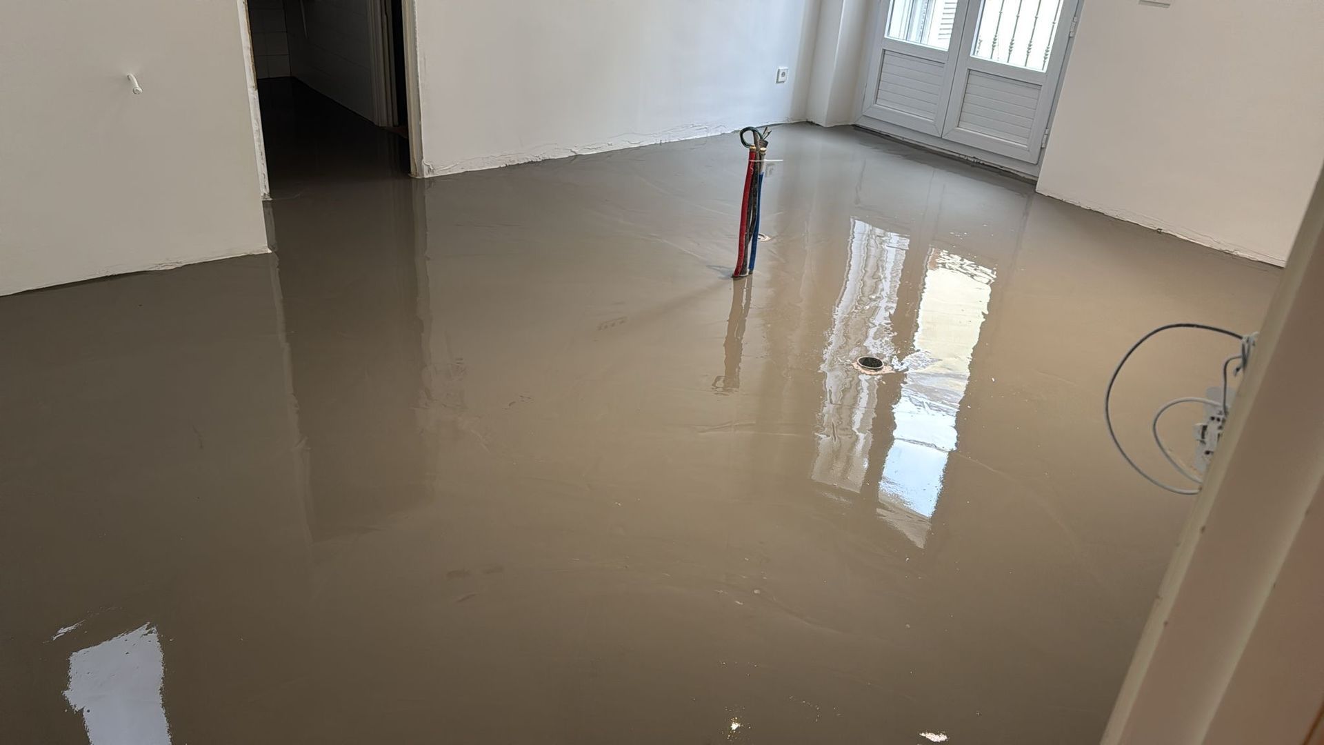 Piso de concreto autonivelante recién vertido y húmedo en una habitación, que refleja la luz desde una ventana y una puerta.