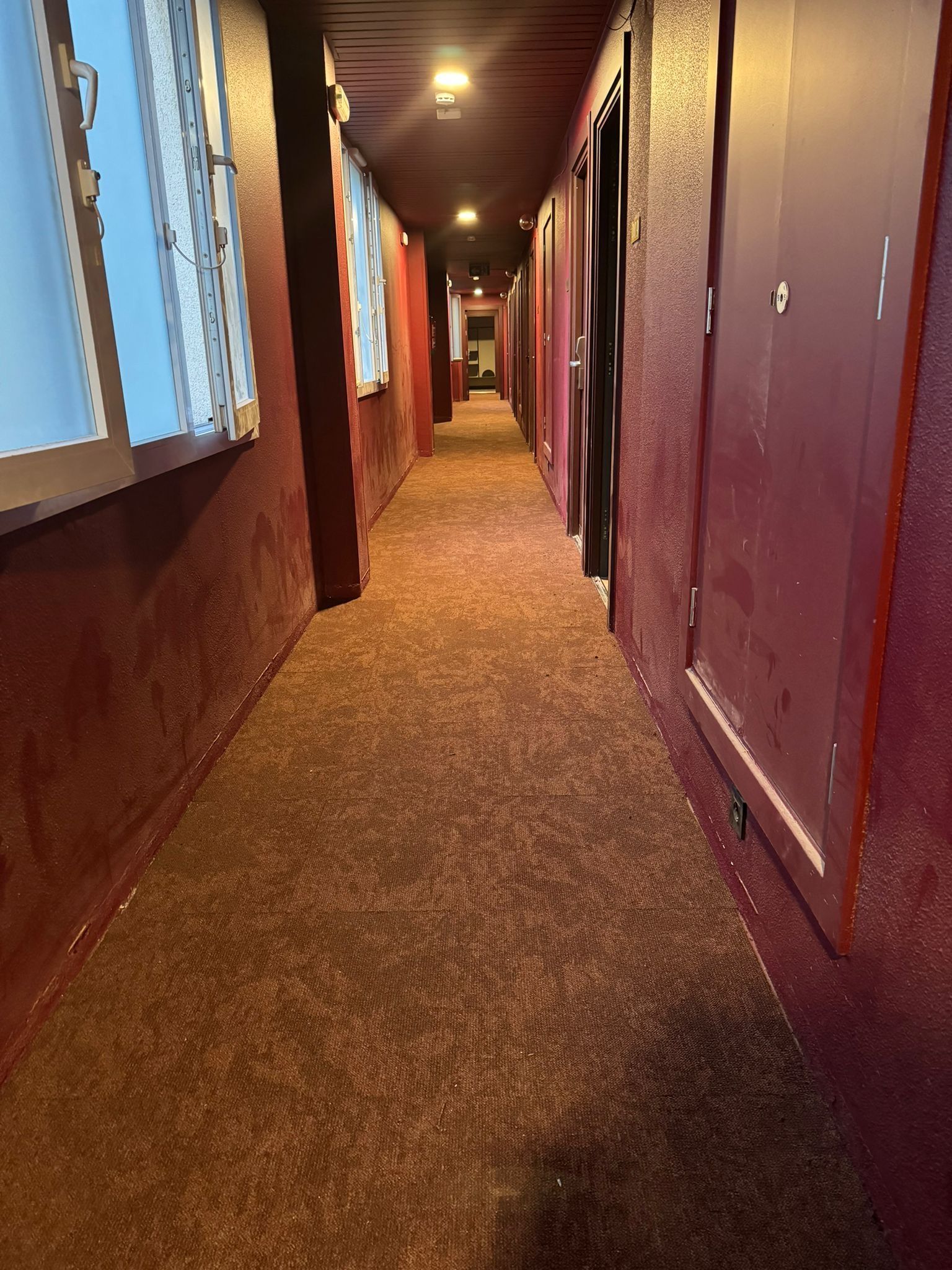 Pasillo estrecho y alfombrado, con puertas a un lado y ventanas al otro, poco iluminado.