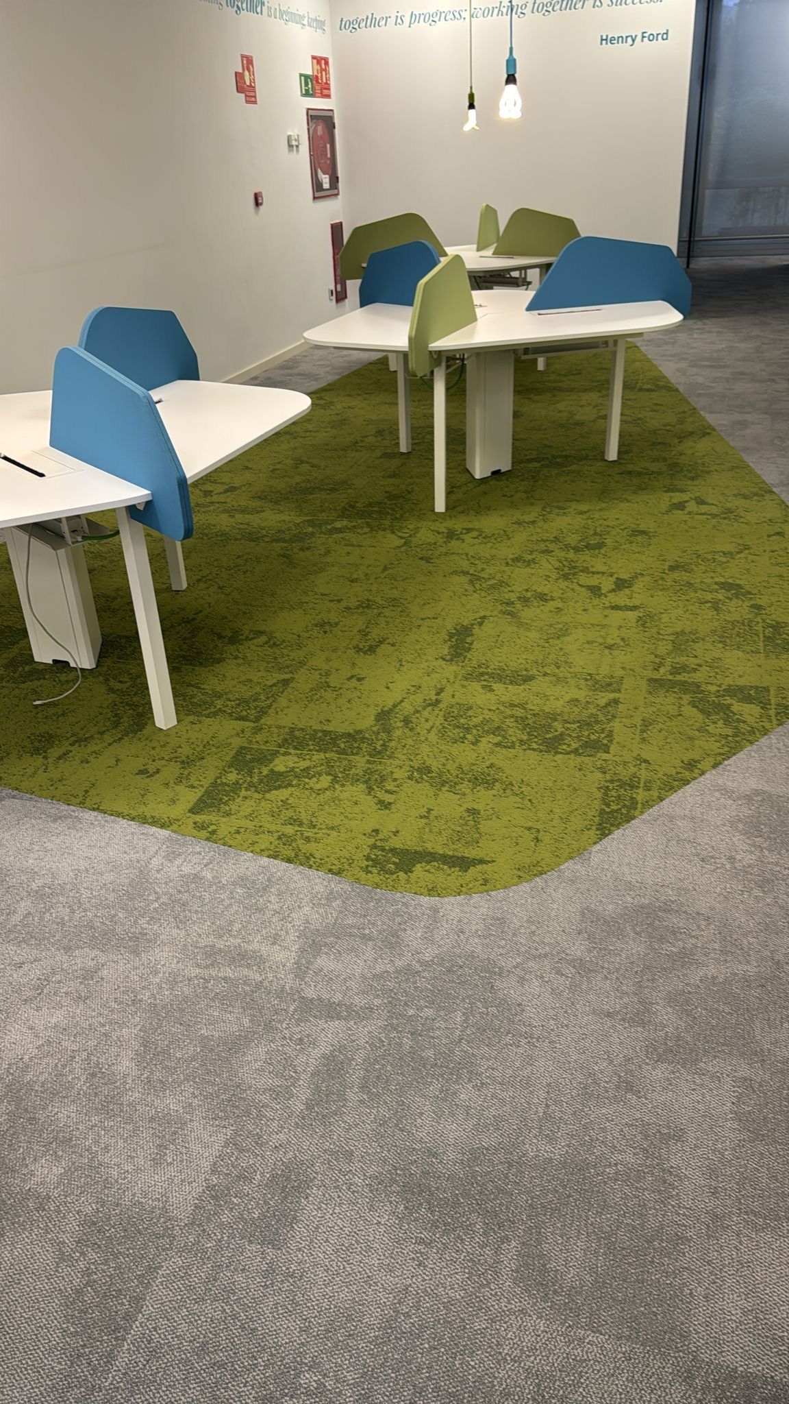 Oficina con área alfombrada verde con escritorios y divisiones azules y verdes. Alfombra gris alrededor.