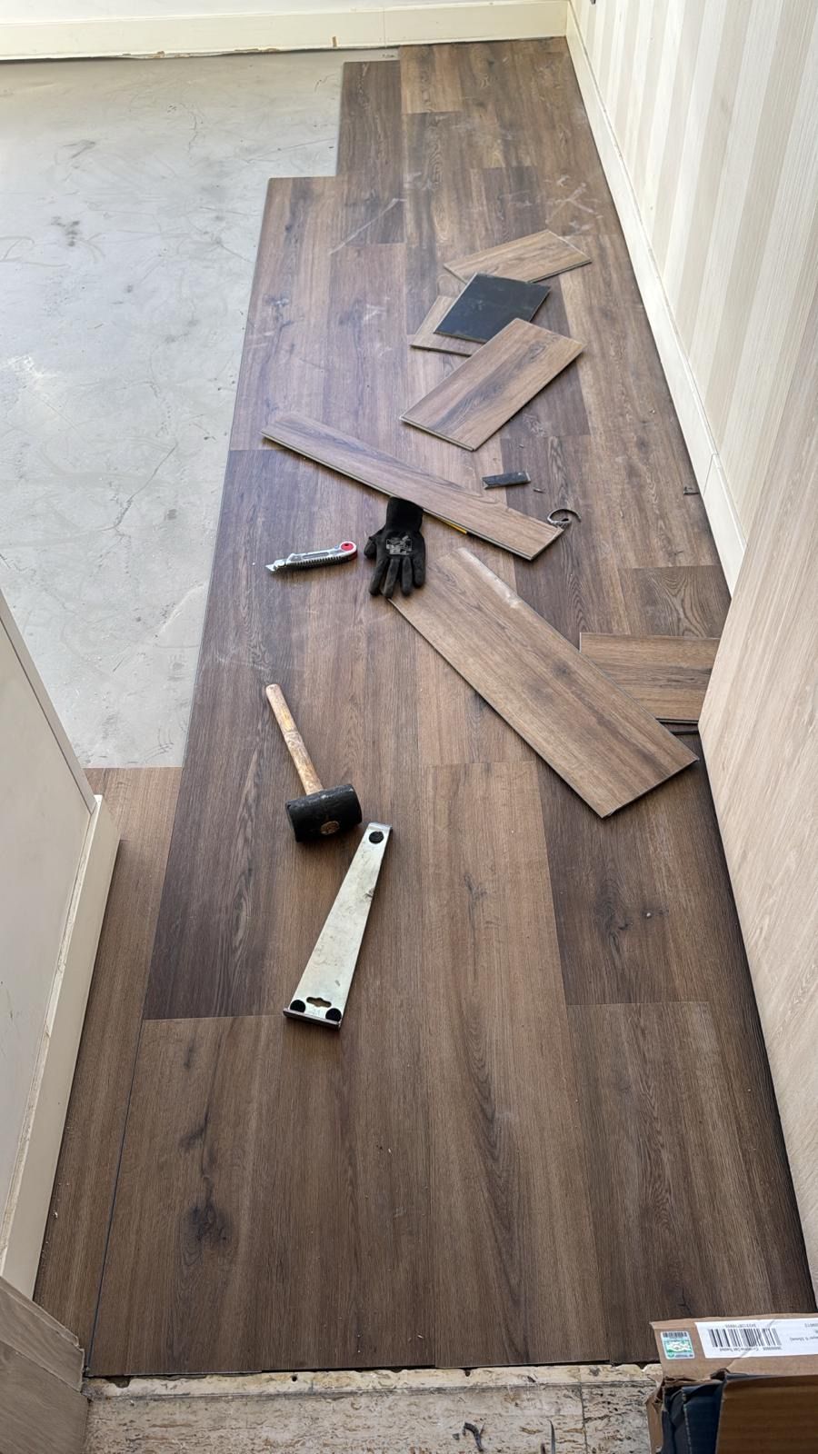 Instalación de suelo marrón imitación madera. Se ven herramientas como un martillo y guantes en el suelo.