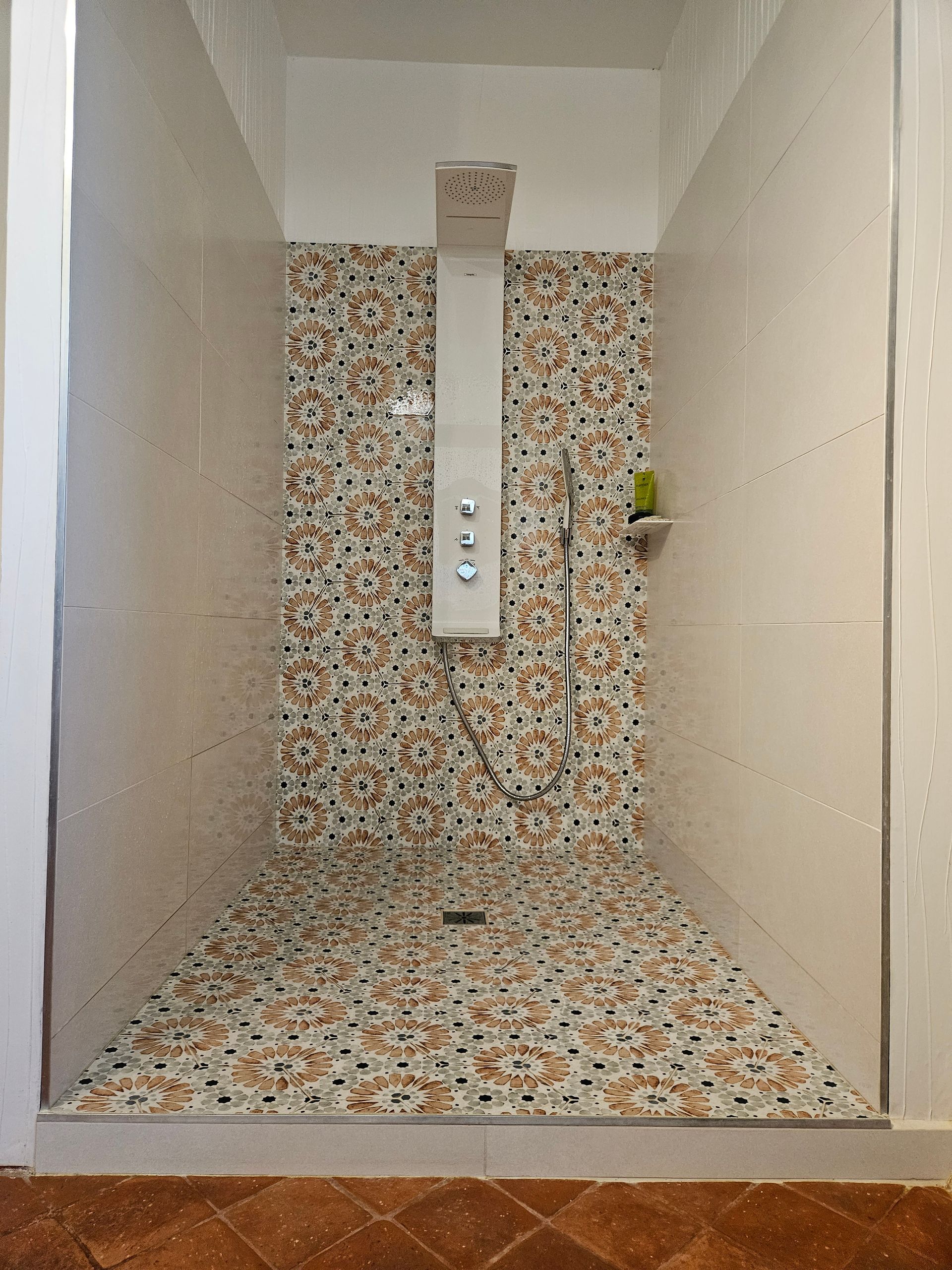 Cabine de douche carrelée vide avec sol et panneau mural en mosaïque florale, plus une petite étagère et une poignée.