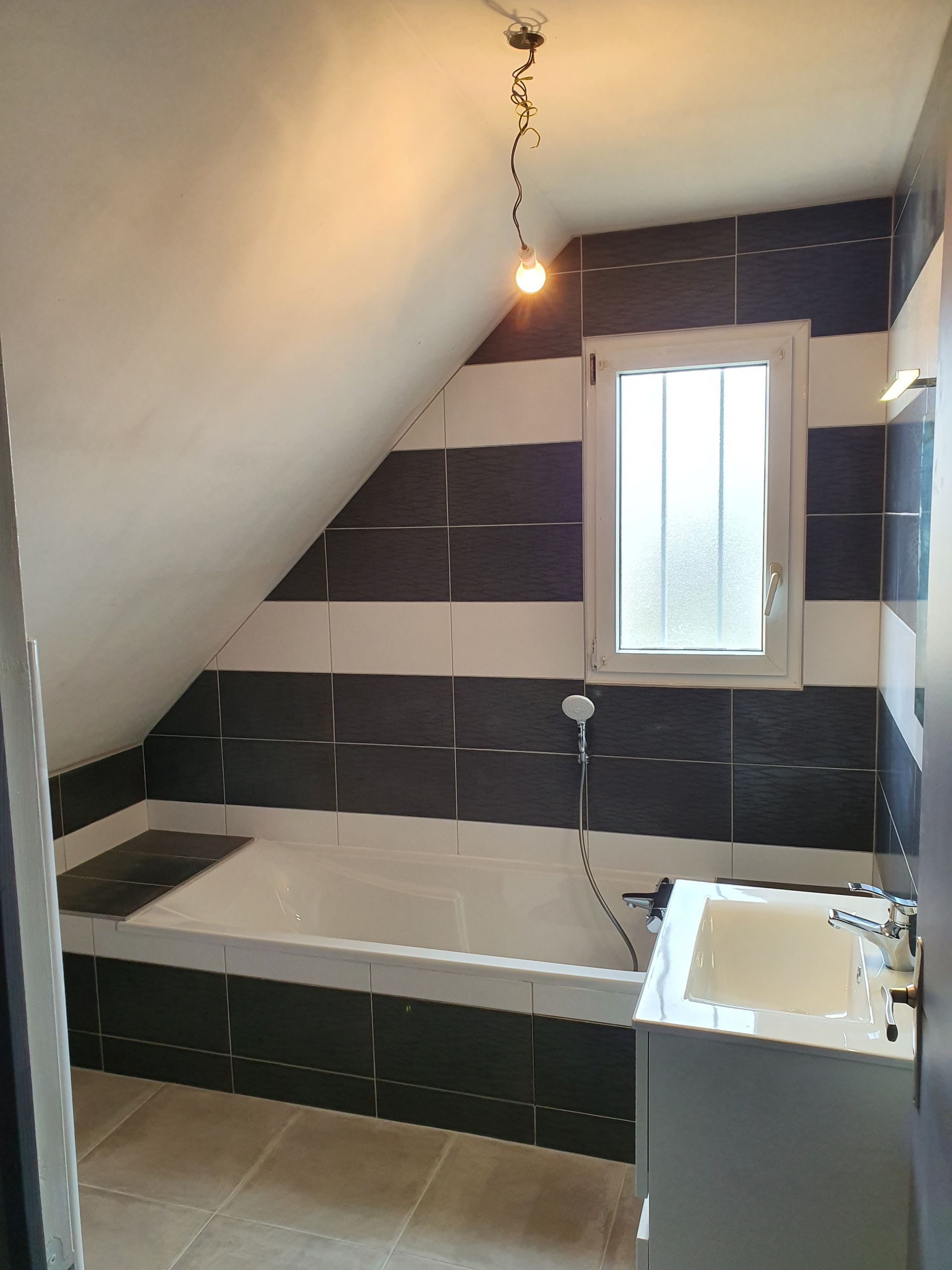 Petite salle de bain mansardée avec murs carrelés, baignoire, lavabo et une fenêtre étroite sous un plafond en pente