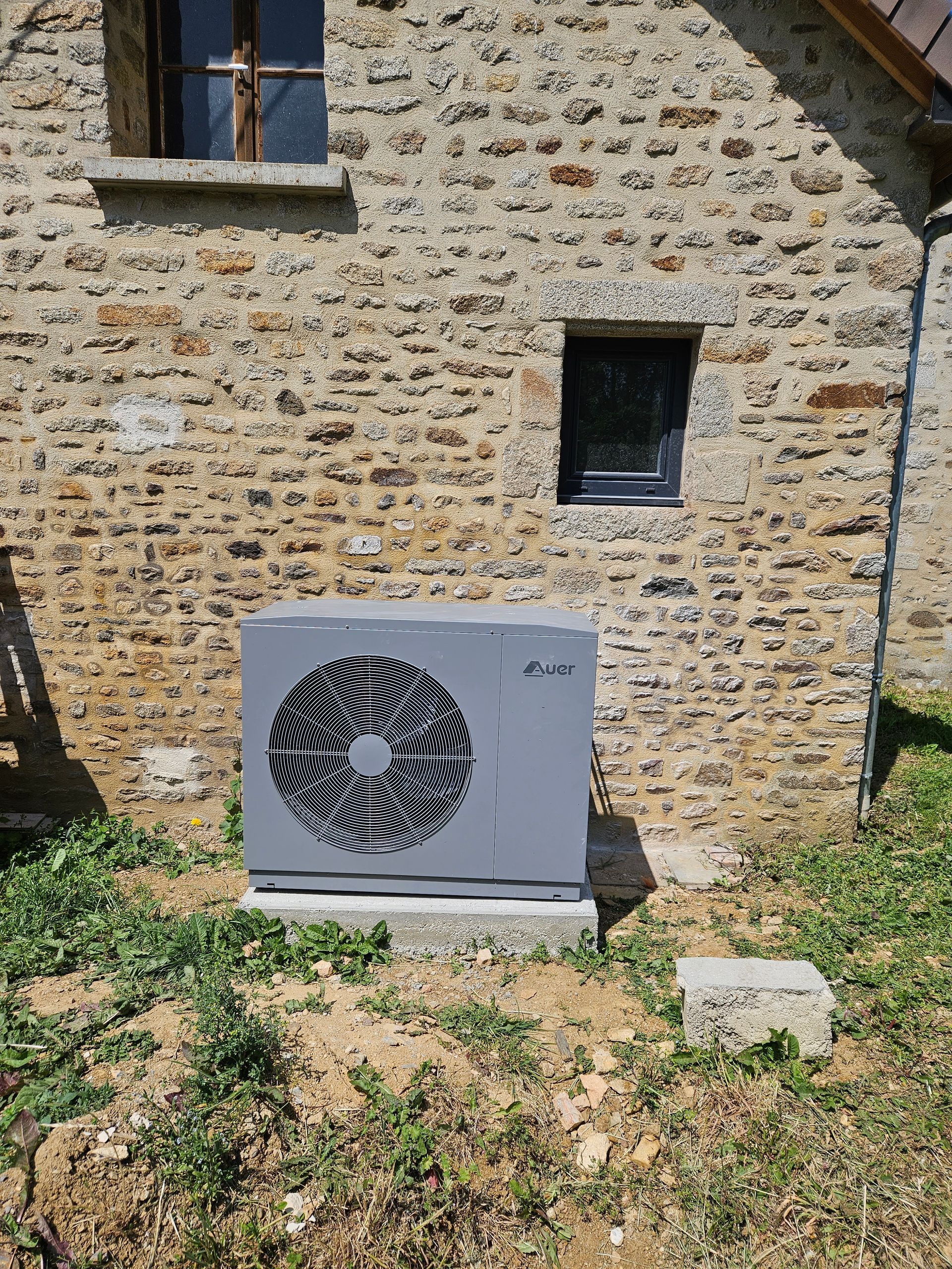 Unité extérieure de pompe à chaleur grise à côté d'un mur en pierre avec deux petites fenêtres et de l'herbe sèche devant