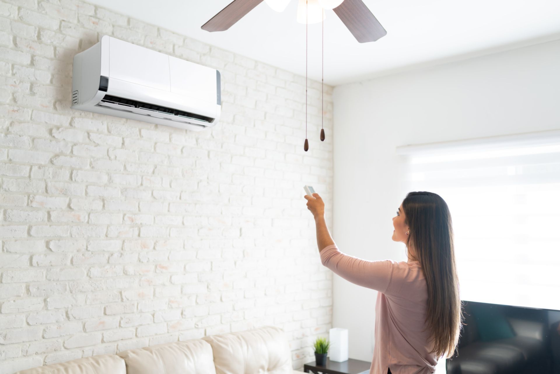 Une femme pointe une télécommande vers un climatiseur mural dans un salon lumineux.