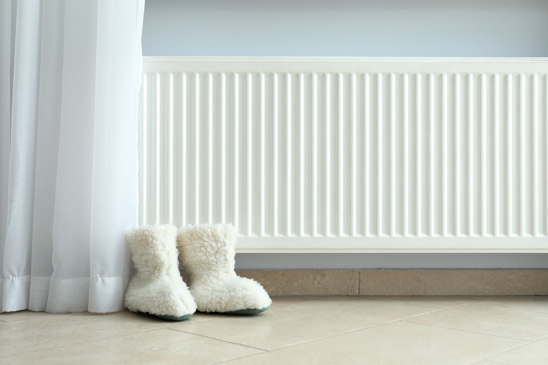 Des bottes blanches duveteuses près d'un rideau transparent, à côté d'un radiateur blanc, dans une pièce minimaliste.