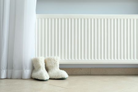 Des bottes blanches duveteuses près d'un rideau transparent, à côté d'un radiateur blanc, dans une pièce minimaliste.