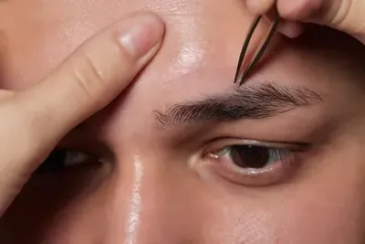 Un primer plano de una persona depilando las cejas de un hombre con pinzas.