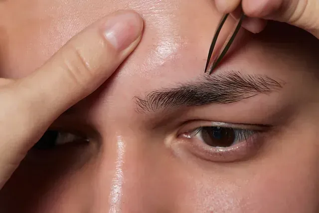 Un primer plano de una persona depilando las cejas de un hombre con pinzas.