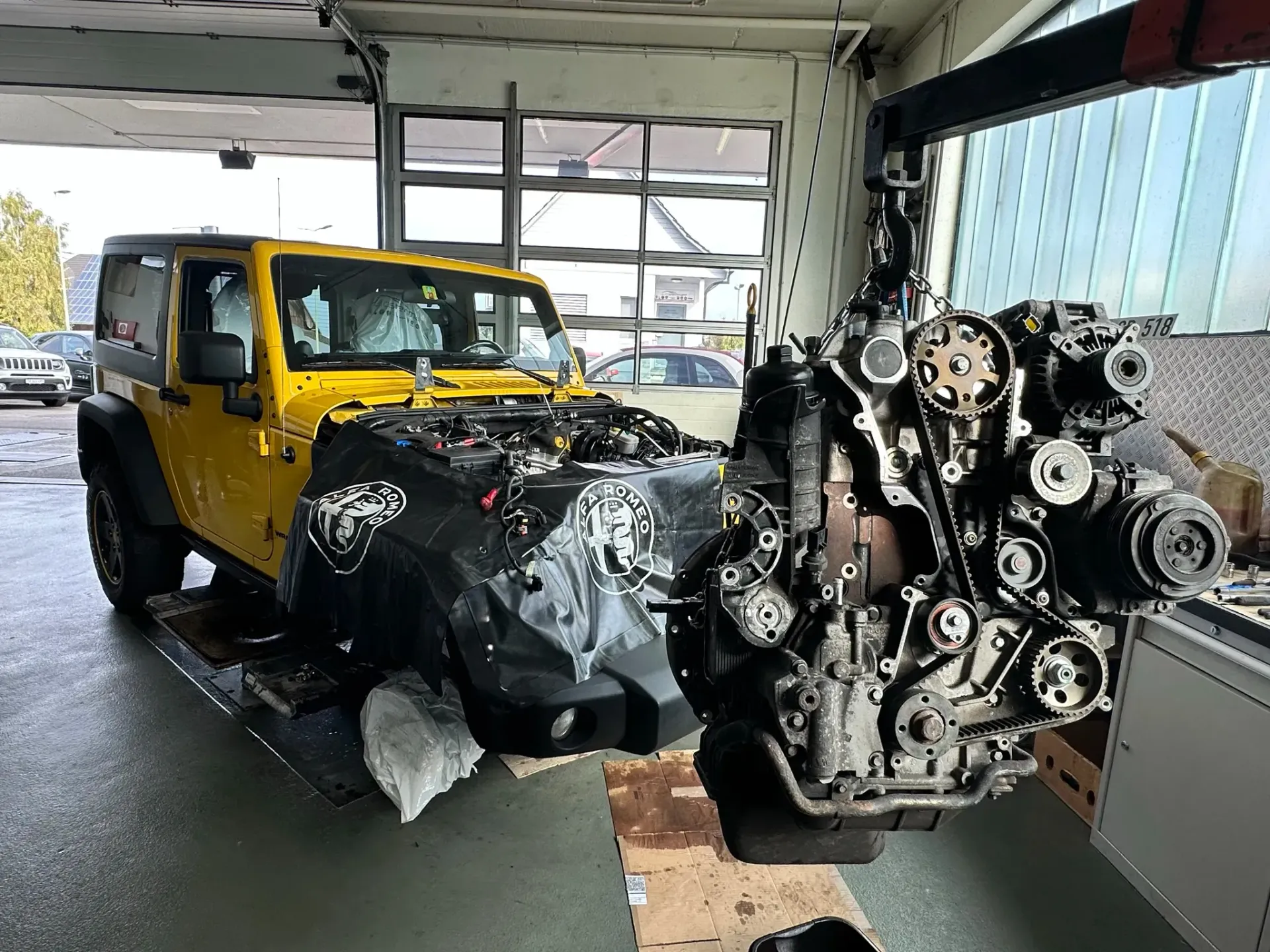 In einer Garage wird an einem gelben Jeep gearbeitet, dessen Motor ausgebaut ist.