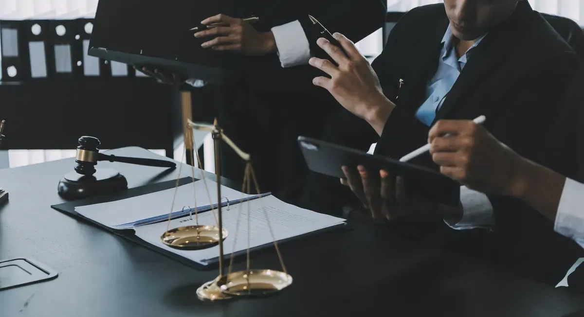 Abogados en una mesa, discutiendo documentos y usando una tableta; el mazo y la balanza de la justicia están en primer plano.
