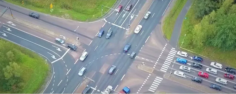 Ilmakuva vilkkaasta risteyksestä, jonka sivuilla on autoja, suojateitä ja rehevää vihreää kasvillisuutta.