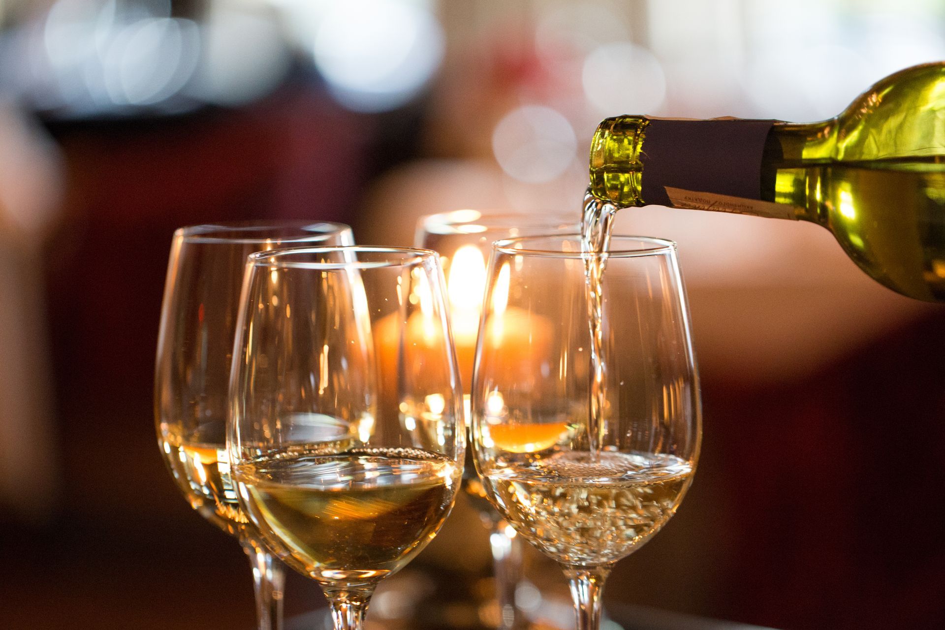 Du vin blanc est versé dans un verre, avec plusieurs verres remplis et des bougies allumées en arrière-plan.