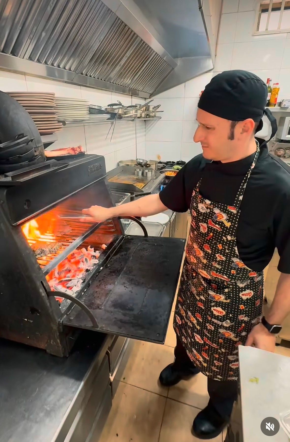 Un chef, con gorro y delantal negros, vigila las llamas de una parrilla en la cocina de un restaurante.