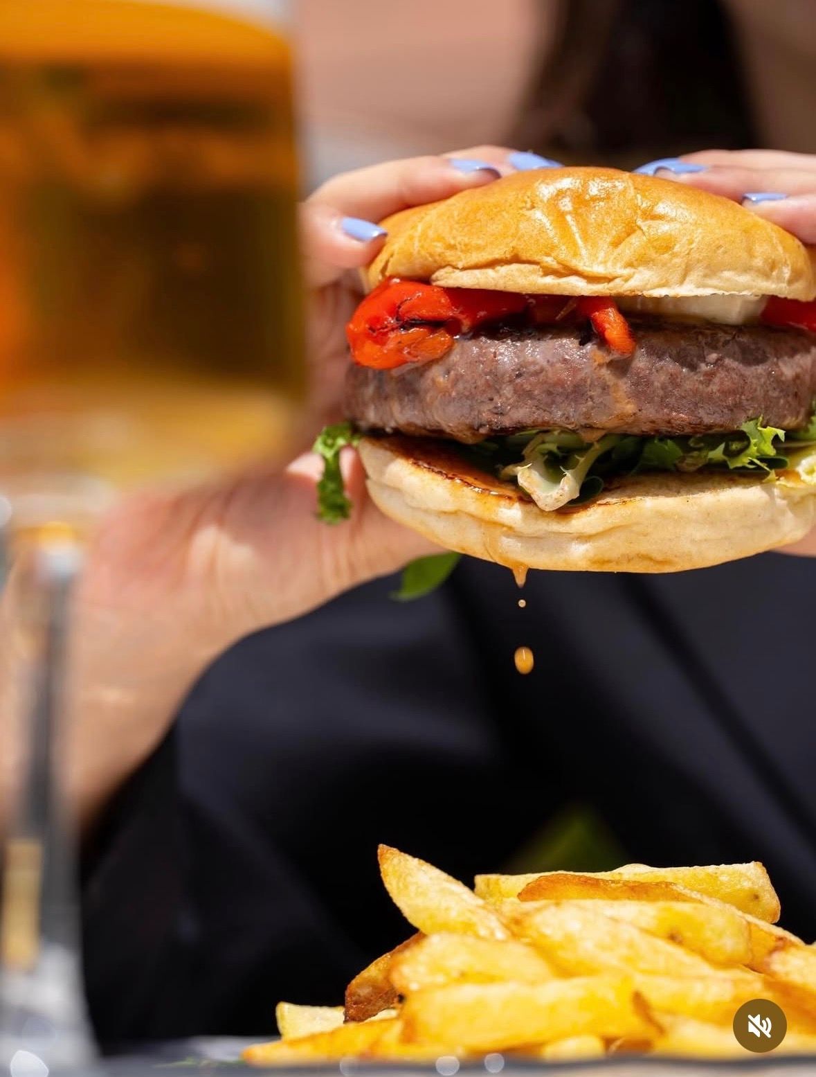 Una mano sosteniendo una hamburguesa con papas fritas en primer plano, con una bebida borrosa al fondo.