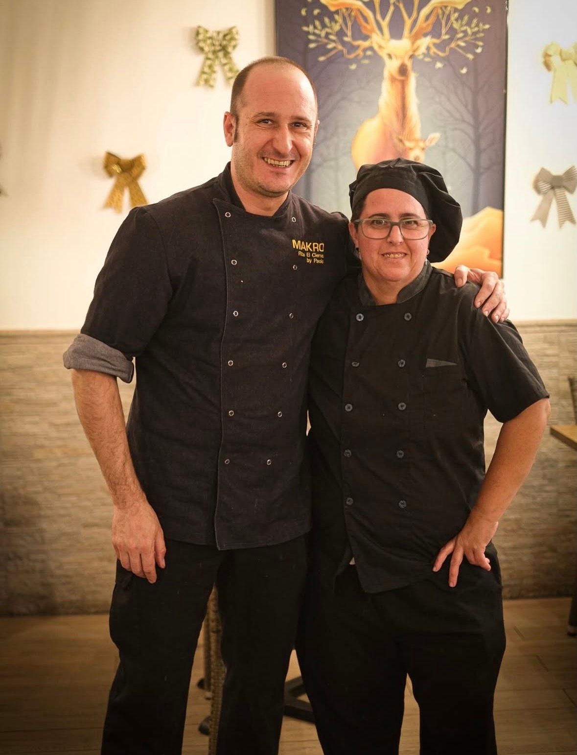 Dos personas con uniformes de chef negros posan juntas en el interior, sonriendo frente a una decoración con forma de ciervo.