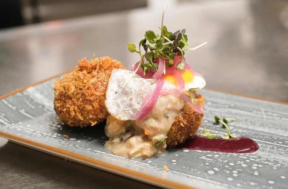 Croquetas gourmet emplatadas con salsa cremosa, microvegetales y sirope de frutos rojos en un plato gris.