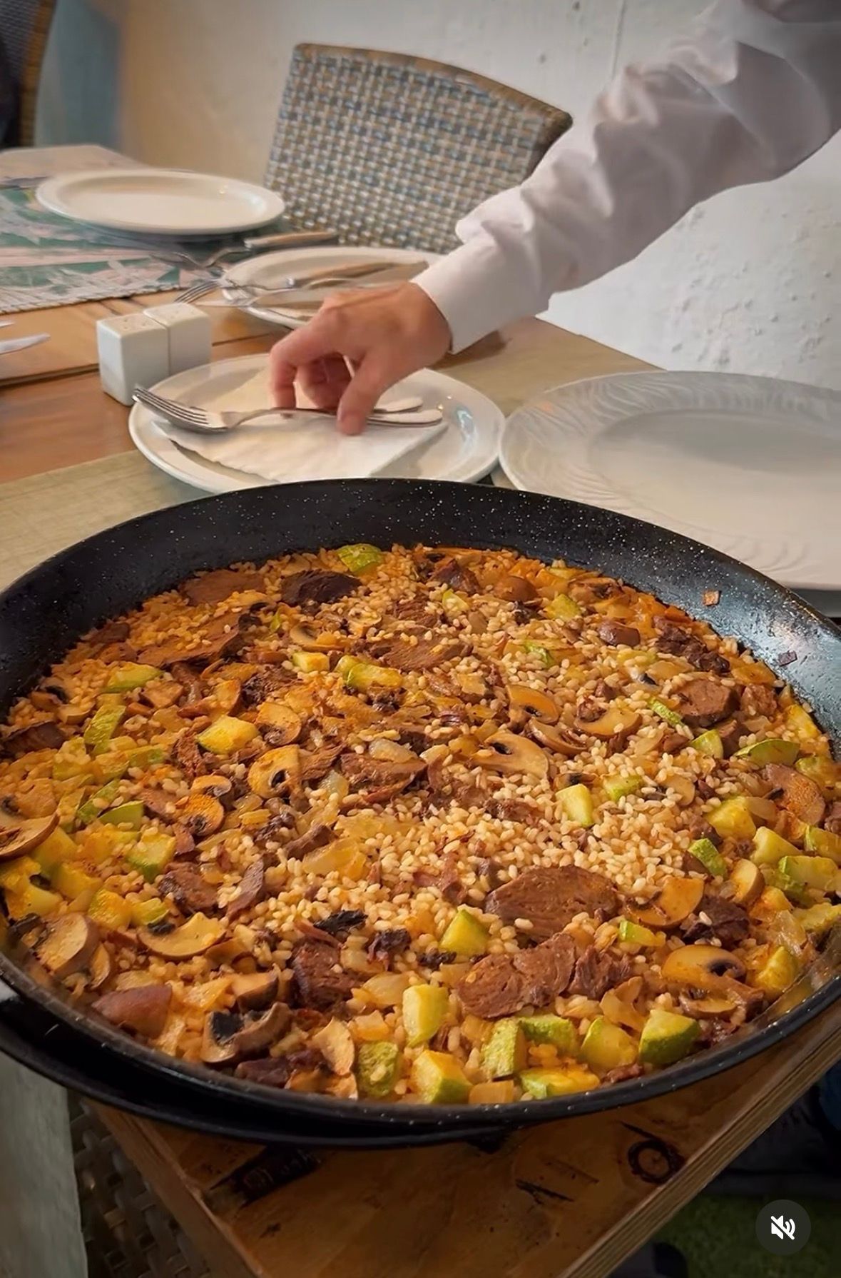 Una mano coloca un plato al lado de una sartén grande con arroz cocido y verduras en una mesa de comedor.