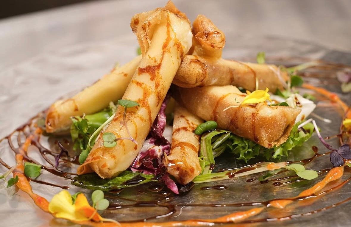 Rollitos fritos emplatados en un plato de cristal, adornados con hierbas y una salsa colorida.