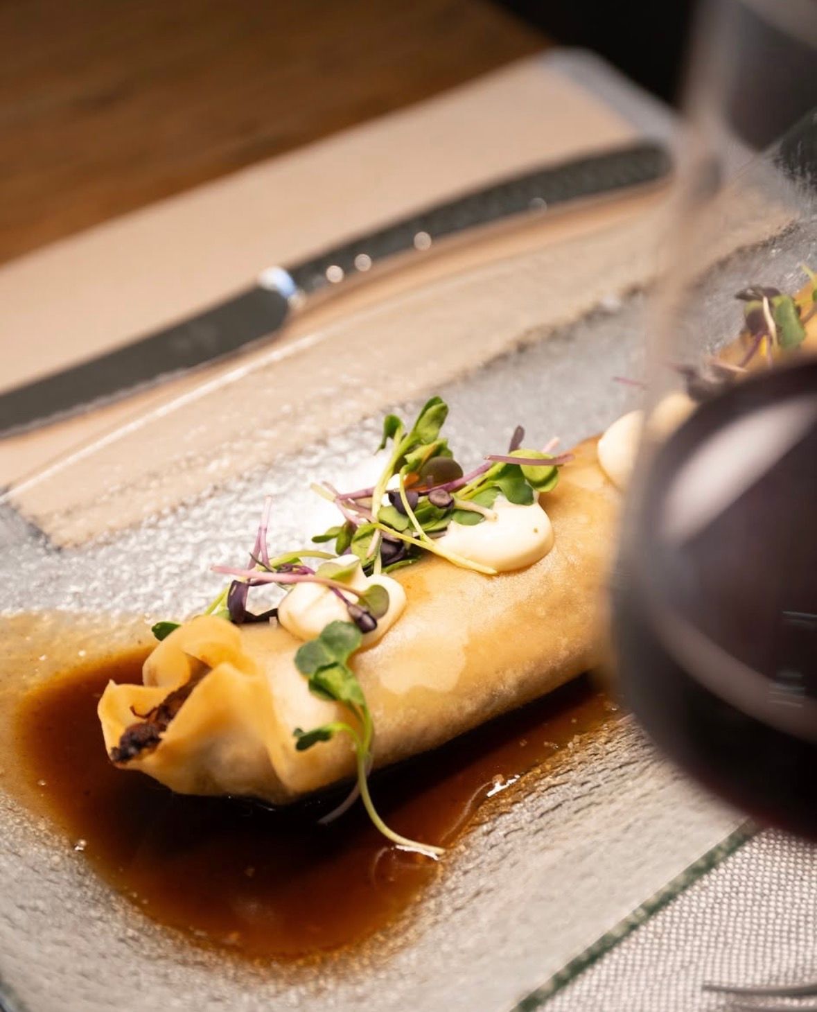 Rollito de primavera emplatado sobre salsa oscura, cubierto con microvegetales y crema blanca, en la mesa de un restaurante.