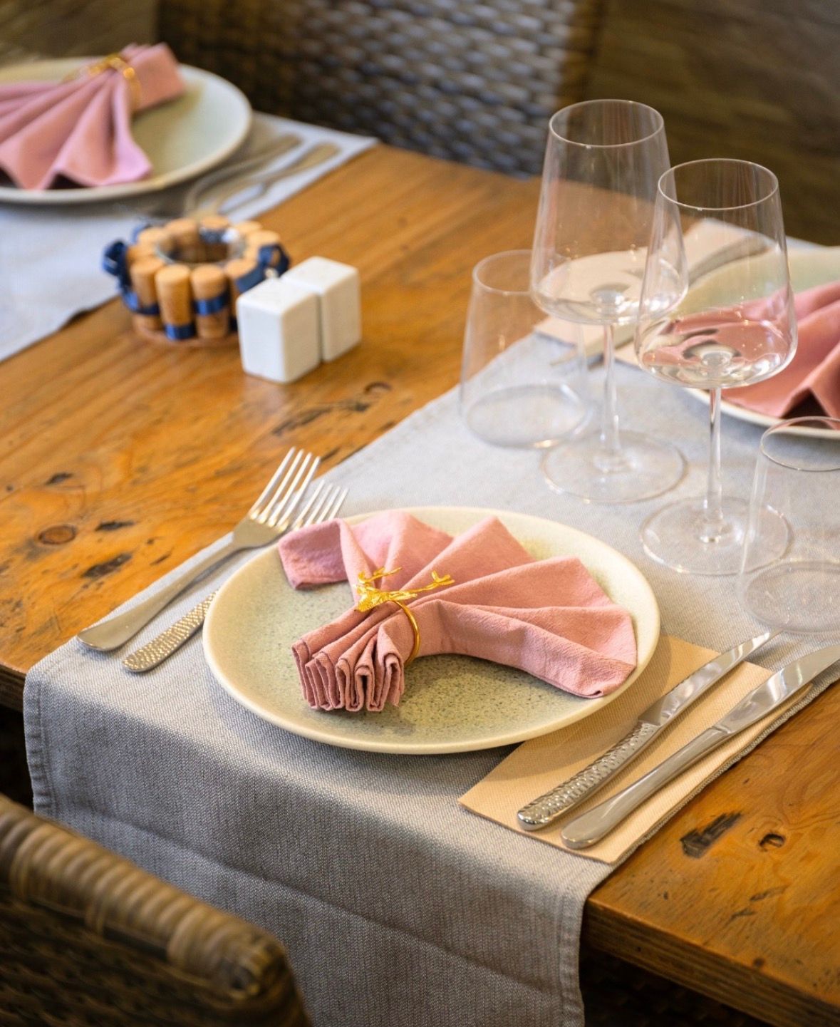 Prepara la mesa de comedor con servilletas rosas, platos blancos, cristalería y sillas de madera sobre una mesa rústica.