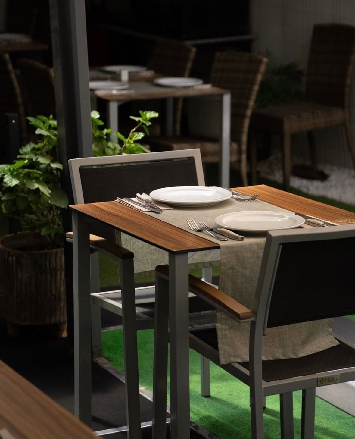 Mesa de café al aire libre con dos platos blancos, tablero de madera y sillas vacías en un patio.