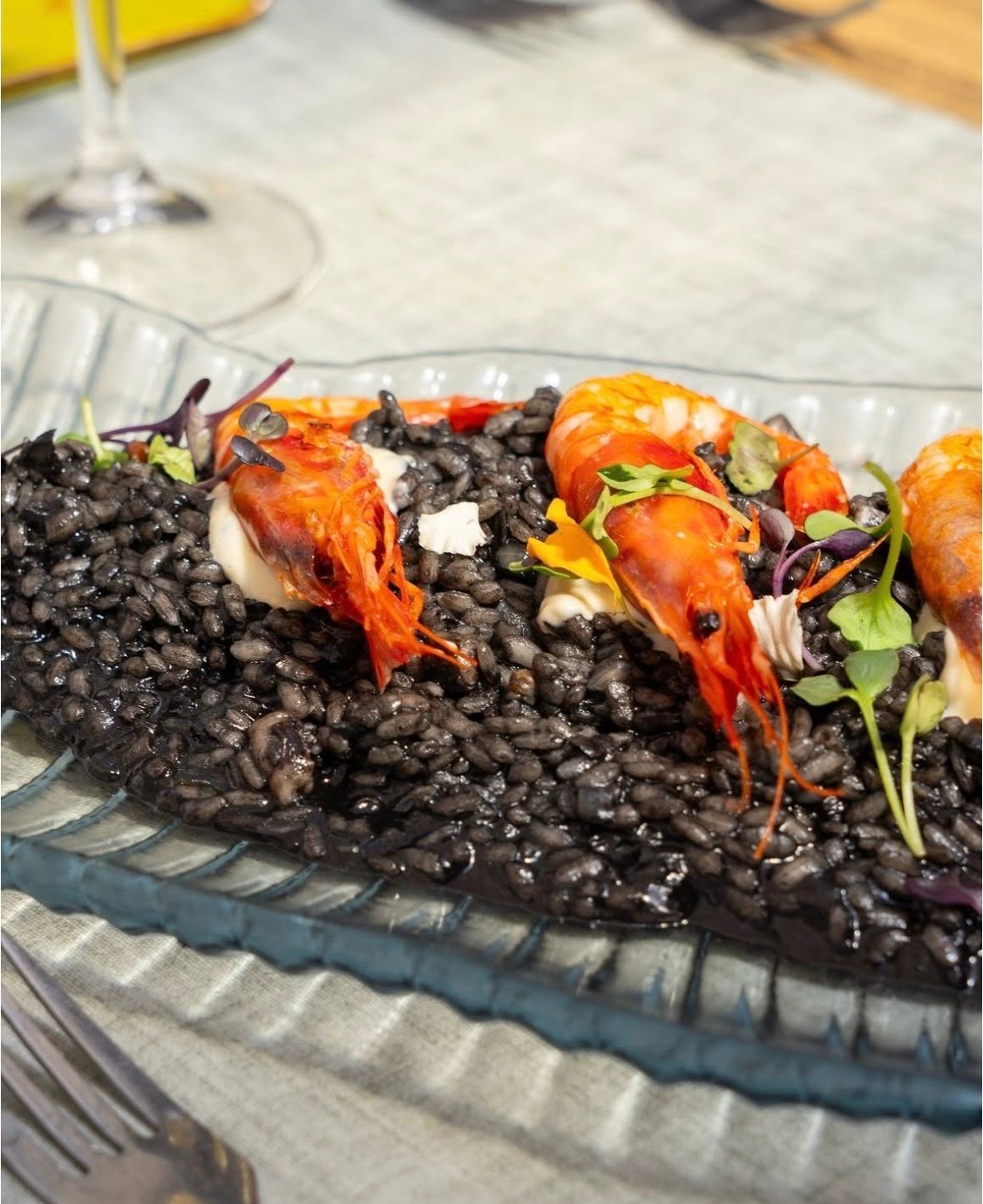 Risotto negro con gambas a la parrilla y guarnición verde en un plato de cristal.