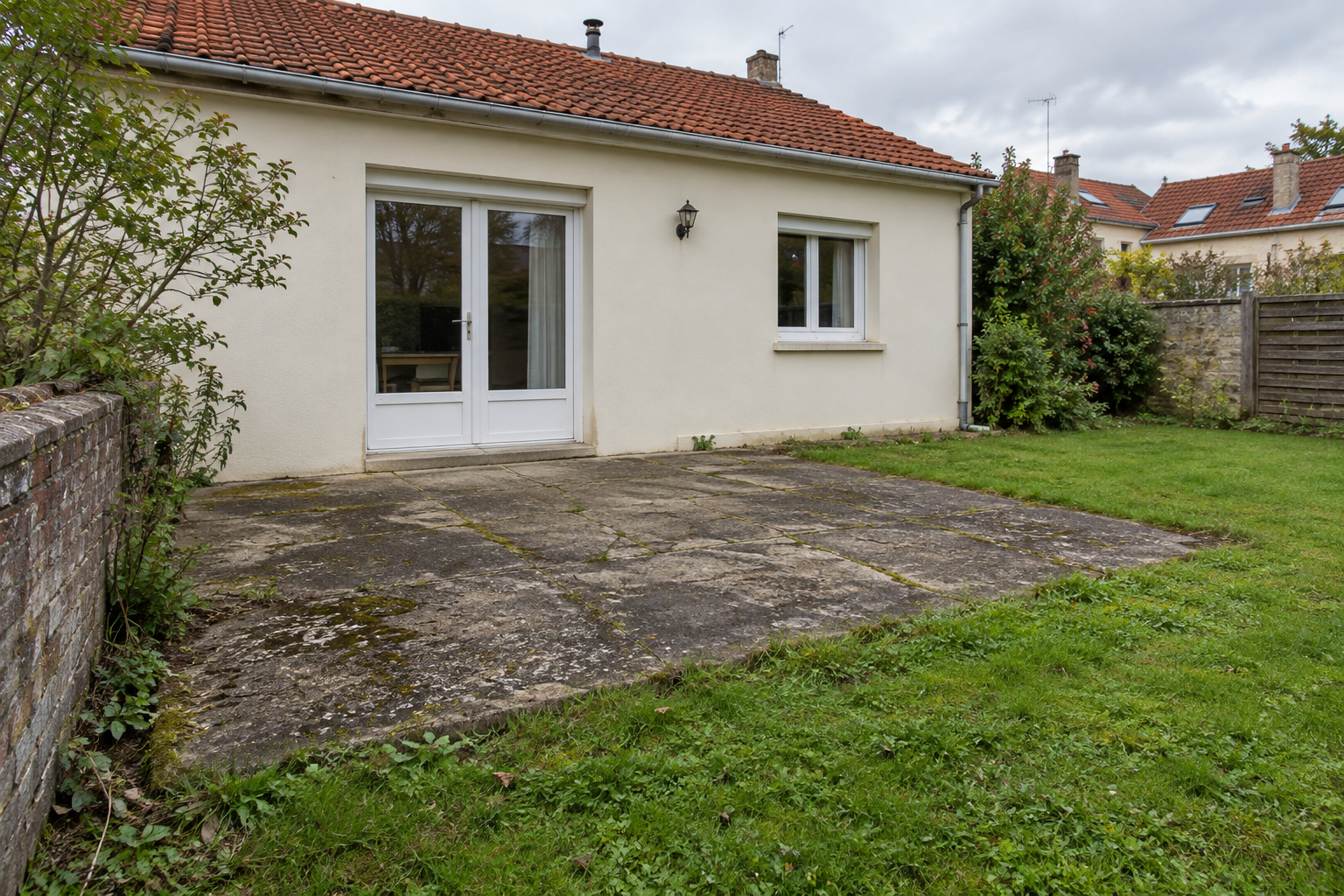 Patio d’une petite maison avec un dallage en pierre, des murs blancs, un toit de tuiles rouges et un jardin gazonné.