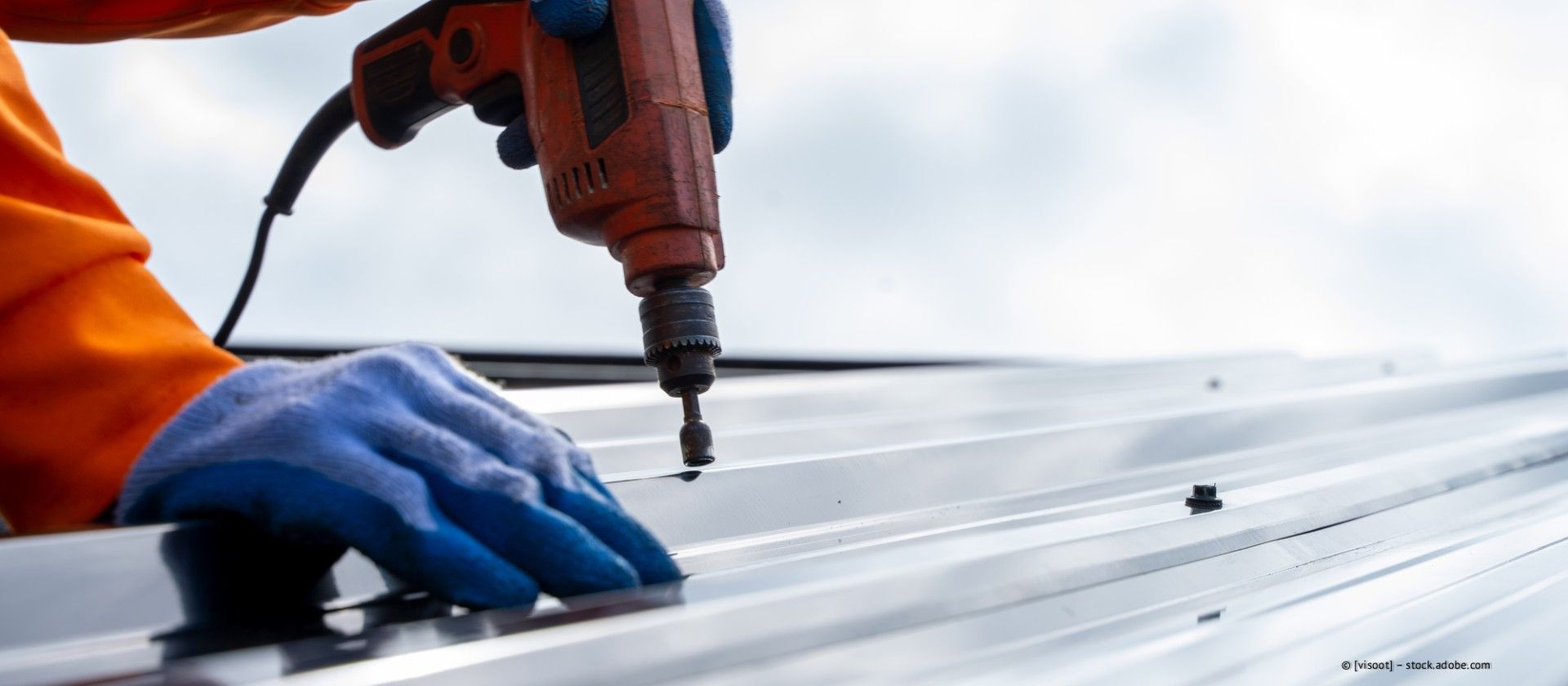 Ein Arbeiter benutzt eine Bohrmaschine auf einem Metalldach und trägt dabei blaue Handschuhe und eine orangefarbene Sicherheitswerkzeugtasche.