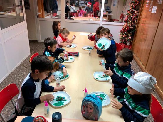 Niños sentados alrededor de una mesa, comiendo en platos en un aula o cafetería luminosa.