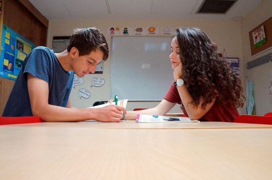 Dos estudiantes escribiendo juntos en una mesa del aula, sonriendo e inclinados sobre su trabajo.
