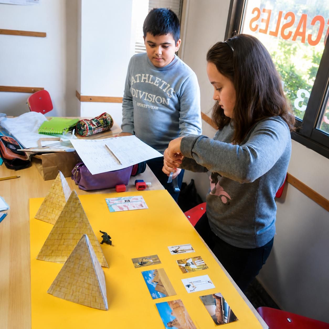 Dos estudiantes trabajando en un proyecto de mesa con maquetas amarillas y papeles en un aula luminosa.