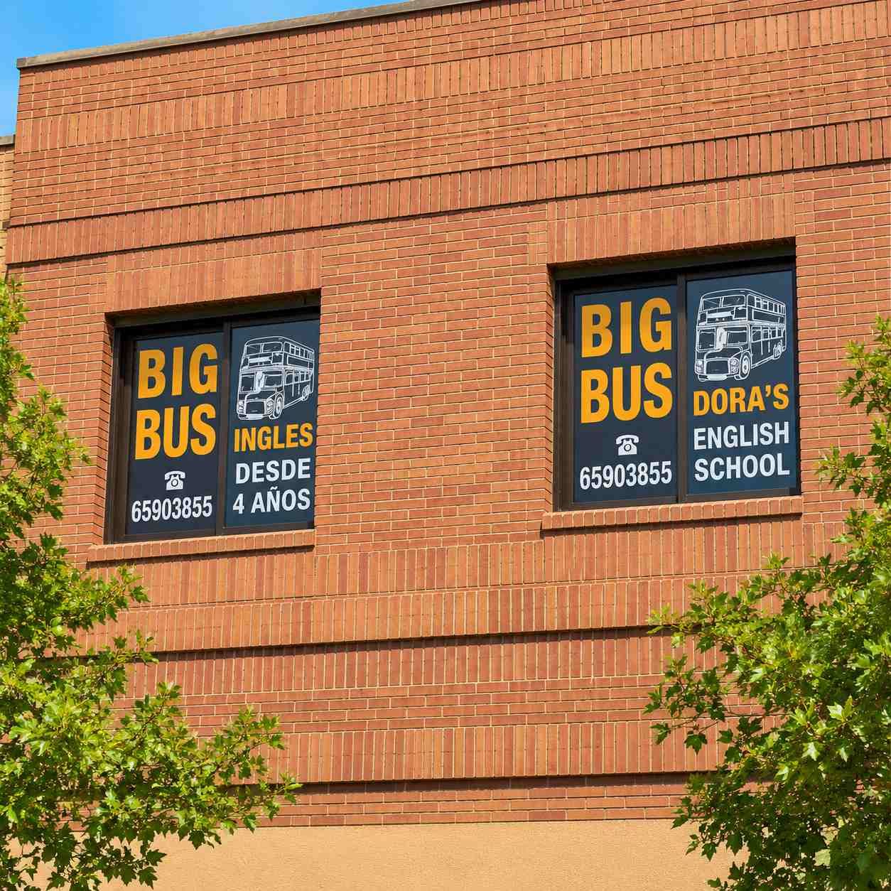 Edificio de ladrillo con dos letreros de Big Bus/Dora's English School en marcos de ventanas negros.