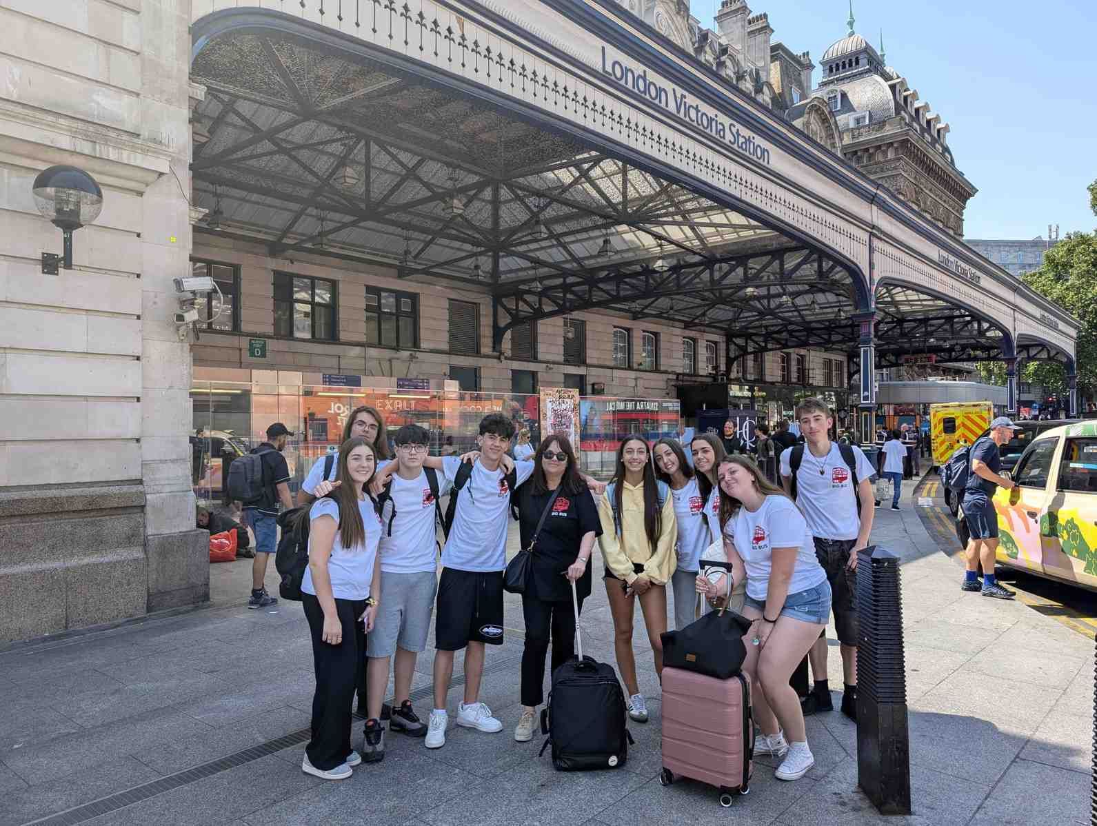 Grupo de viajeros con equipaje posando frente a una estación de tren bajo una gran marquesina.