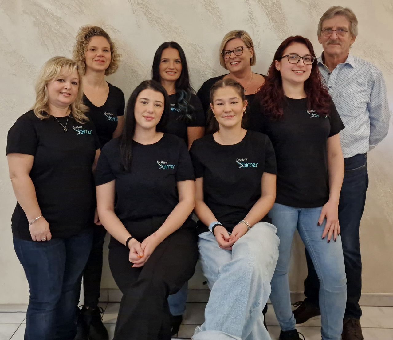 Eine Gruppe von neun Personen in schwarzen Hemden posiert im Innenbereich vor einer neutralen Wand.