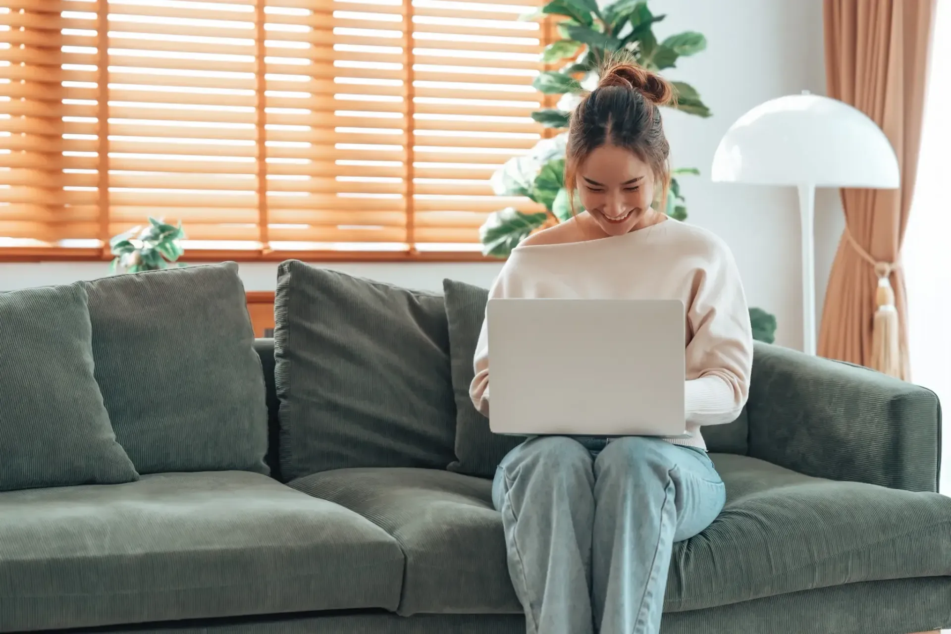 Una mujer sonríe mientras usa una computadora portátil en un sofá verde en una sala de estar.