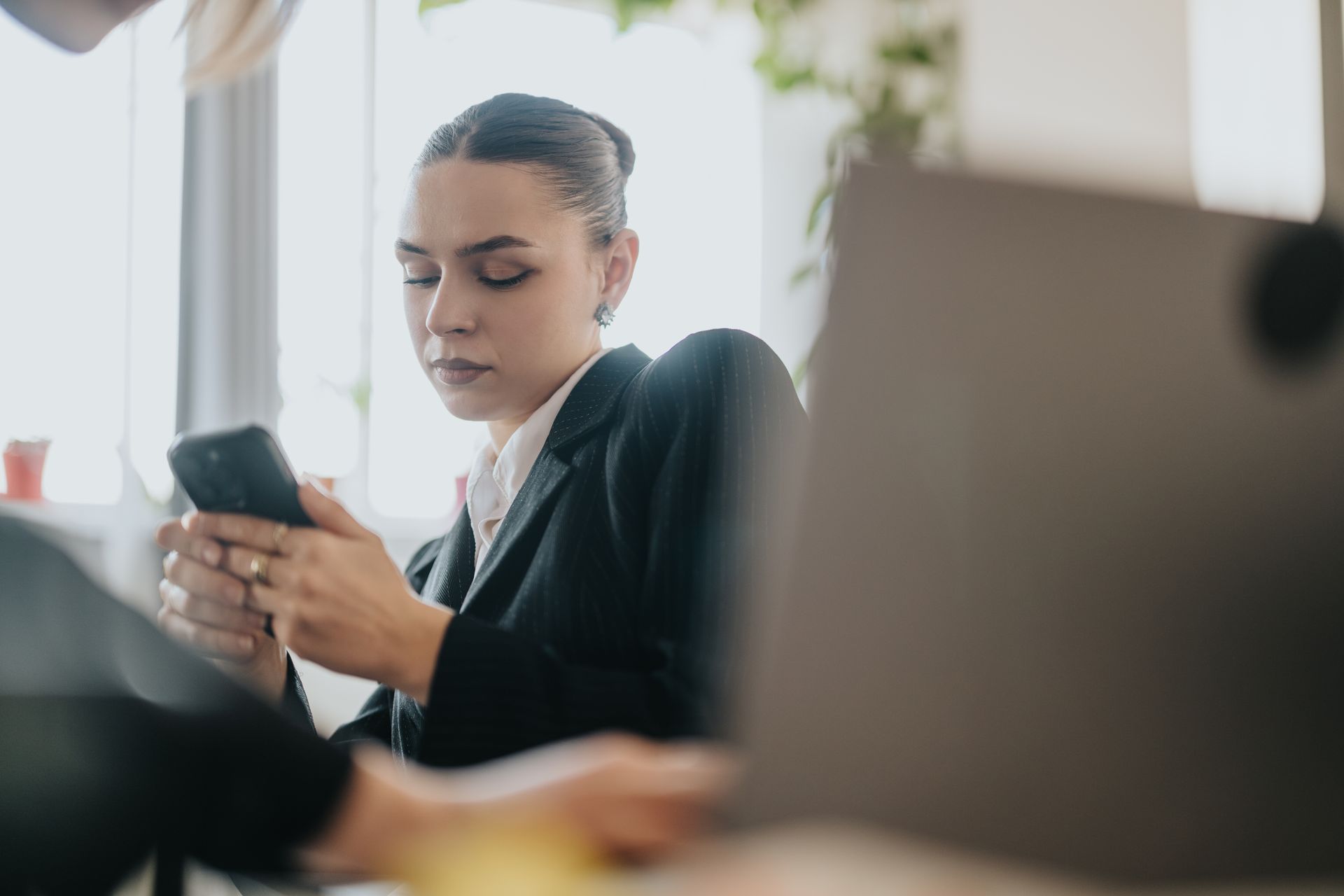 Mujer con un blazer negro mirando su teléfono, sentada cerca de una computadora portátil junto a una ventana.