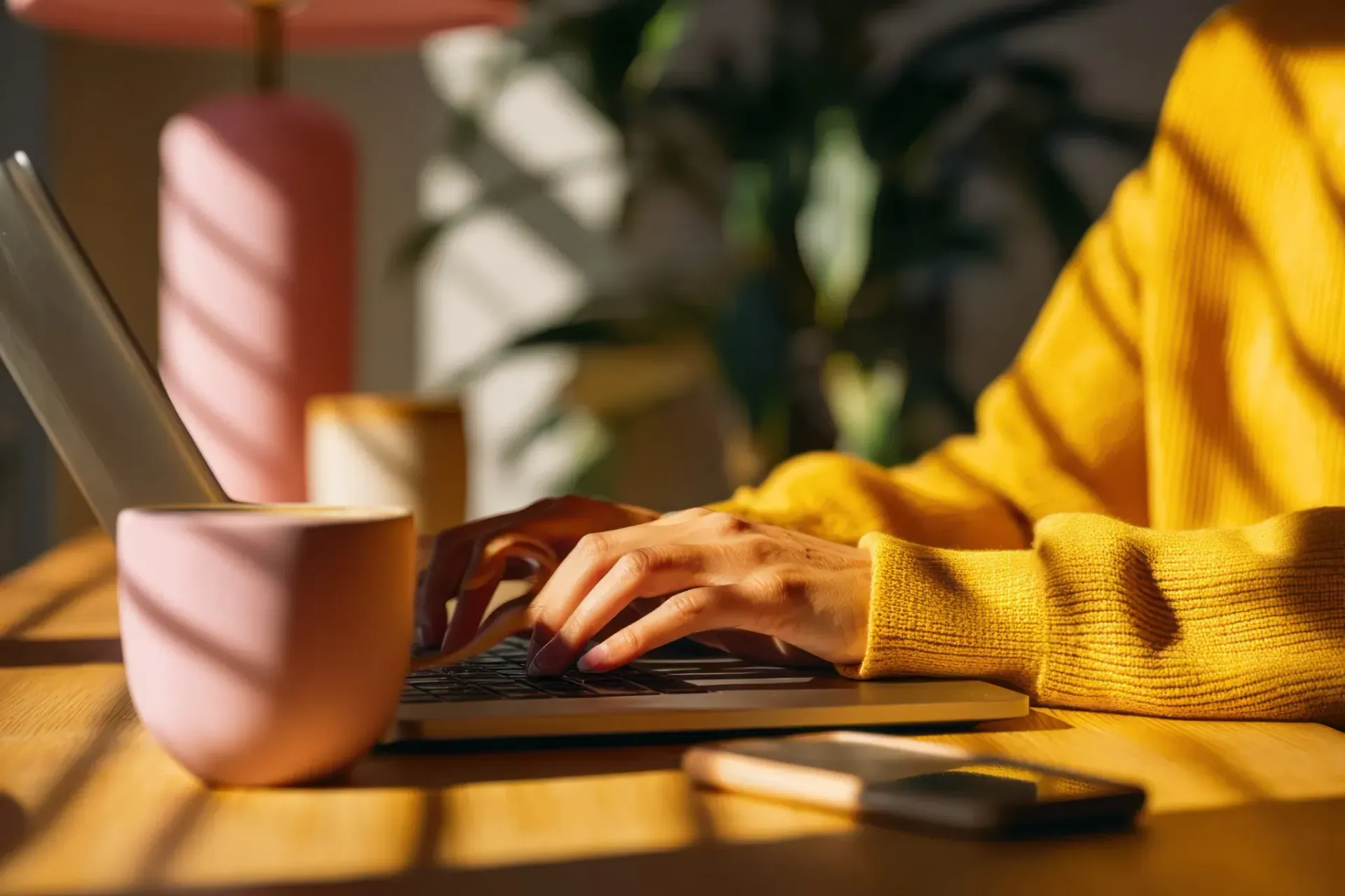 Manos escribiendo en una computadora portátil, vistiendo un suéter amarillo, cerca de una taza rosa y una lámpara.