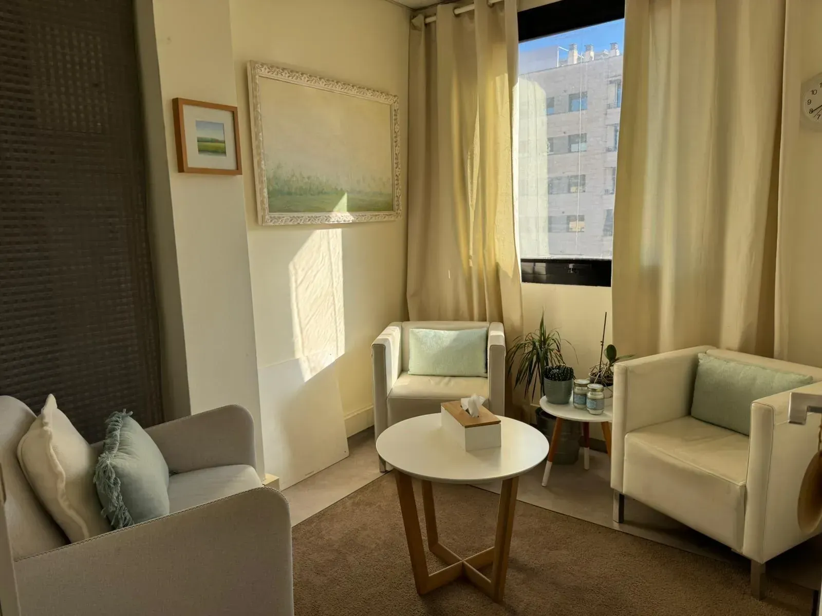 Acogedor consultorio de terapia con sillones de colores claros, pequeña mesa redonda y una ventana con cortinas.
