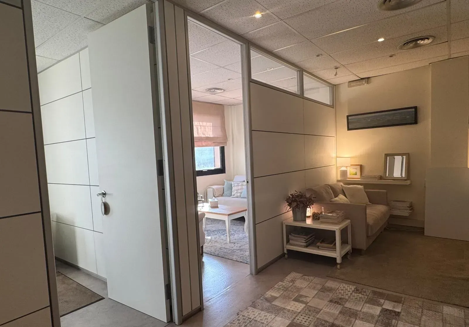 Un pasillo conduce a una habitación con sofá, mesa y ventana. Paredes blancas y luz natural.