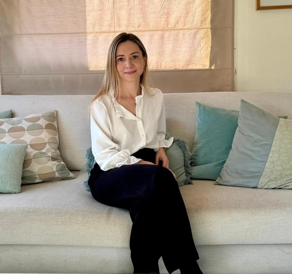 Mujer sentada en un sofá, con camisa blanca y pantalón negro. Luz del sol y cojines azules.