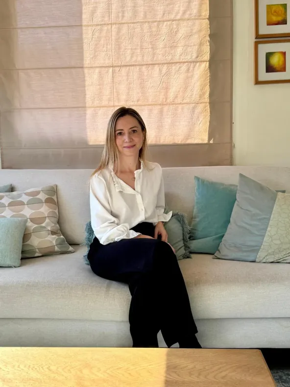 Una mujer con camisa blanca y pantalón negro está sentada en un sofá color crema, mirando a la cámara. Luz tenue y cojines.